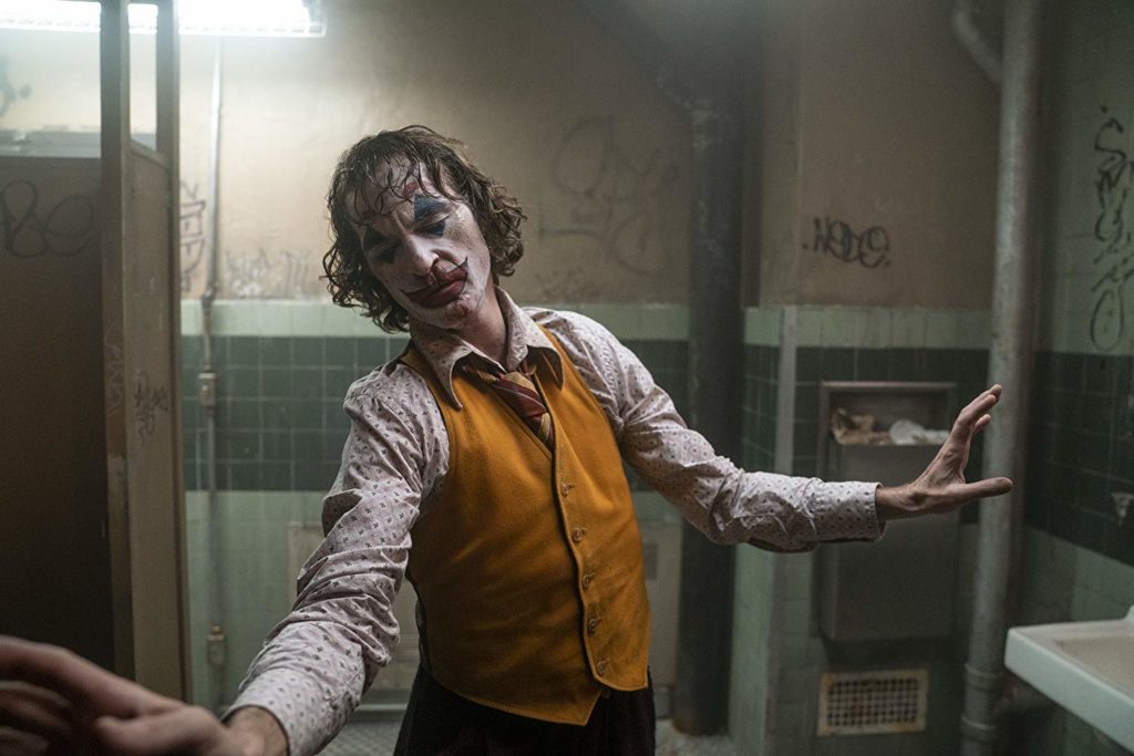 Cena em que o personagem parece executar uma dança num banheiro público. Seus braços estão abertos e buscam a direção da tela e da parede à sua esquerda. seus olhos estão fechados e ele parece estar refletindo, ou prestando a tenção. Ele usa um colete amarelo, uma camisa estampada uma gravata vermelha e amarela, e seus cabelos estão desgrenhados e molhados. Ele está maquiado e nota-se sangue em seu nariz e queixo.