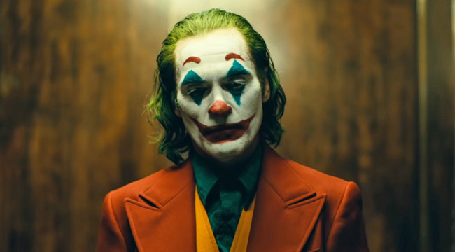 Cena de close no busto do coringa, num plano frontal em que ele olha para baixo da câmera com um olhar semi-cerrado. Mantém uma expressão calma e sua maquiagem contrasta com sua expressão, desenhando um sorriso em seu rosto. Seu cabelo está verde, e ele veste um terno vermelho, um colete amarelo e uma camisa azul. Atrás do personagem uma parede de madeira formada por longos blocos.