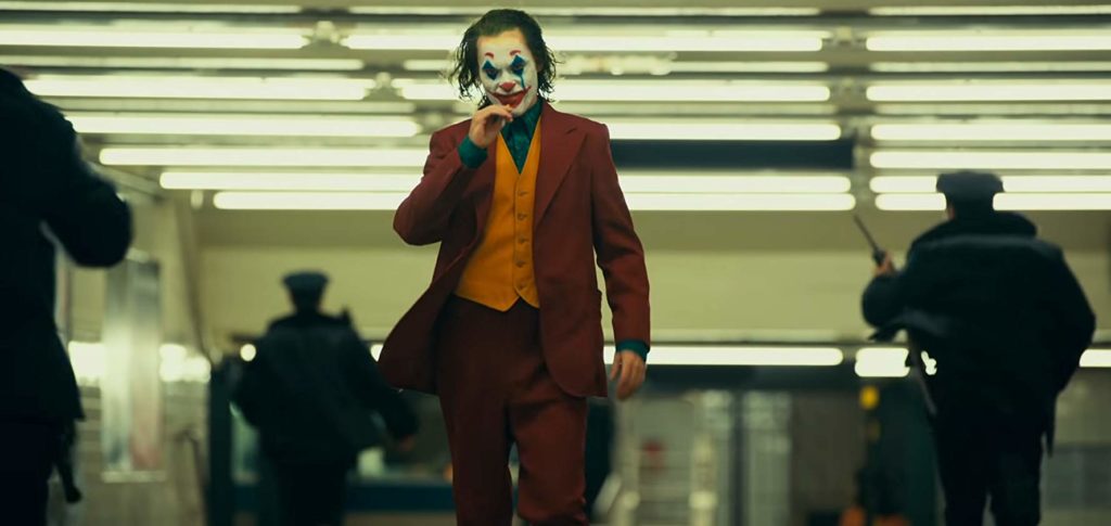 O personagem passa por um salão iluminado por muitas lâmpadas, que parece um terminal de metrô. Ele está vestido de coringa, com terno vermelho, colete amarelo e camisa azul. Seus cabelos estão molhados e a maquiagem ligeiramente borrada. uma mancha risca seu rosto partindo da bochecha esquerda. Ele tem uma expressão confiante. Ao seu redor três policiais correm na direção oposta a dele, 2 já tendo o ultrapassado, indo para um ponto além do que está registrado na cena.