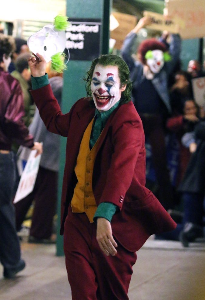 O personagem, vestido de coringa, segura uma máscara em sua mão direita, enquanto parece dançar, com um riso de boca aberta e olhos fechados. Atrás do coringa, uma pessoa está usando a mesma máscara, e levantando um cartaz, e outras parecem parte de um protesto. Em uma pilastra atrás do personagem está uma placa onde se lê "Bedford Park Blvd"
