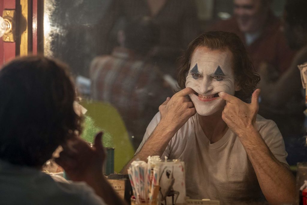 Outra cena em que o personagem, maquiado como palhaço, força um sorriso com os dedos na boca. Nessa cena ele se encontra de frente pro espelho, vestindo uma camisa branca, e vemos a cena de trás do personagem, enxergando no canto direito da tela suas costas e vendo sua face através do espelho. A cena se passa no que parece um camarim, com alguns objetos espalhados na frente do espelho, como um copo com pinceís, um retrato, entre outros não identificáveis.
