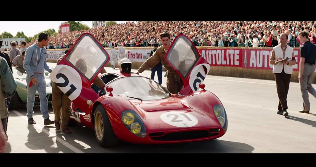 Cena em que o carro da ferrari aparece em destaque, com dois homens empurrando o carro de portas abertas. O carro é numerado como 21. Atrás desse carros vem um carro azul. 