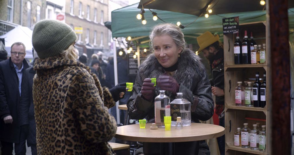 Kate e Petra, numa espécie de feira livre, experimentam uma bebida. Kate está de costas para o leitor, Petra de frente, olhando para ela Parece tímida com seu copo de dose na mão.