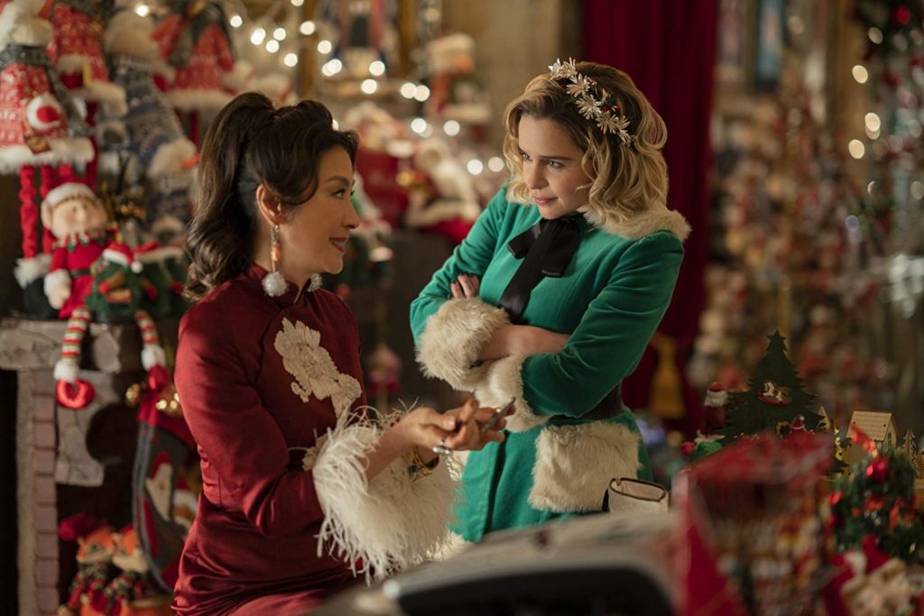 Kate olha para Noel com olhar julgador e divertido, enquanto Noel sorri. estão dentro da loja (portanto, decorações de natal mil ao fundo)