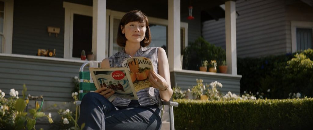 Cena em que Mollie Miles senta no jardim de sua casa, com uma revista aberta, enquanto algo chama sua atenção e ela faz uma expressão de diversão