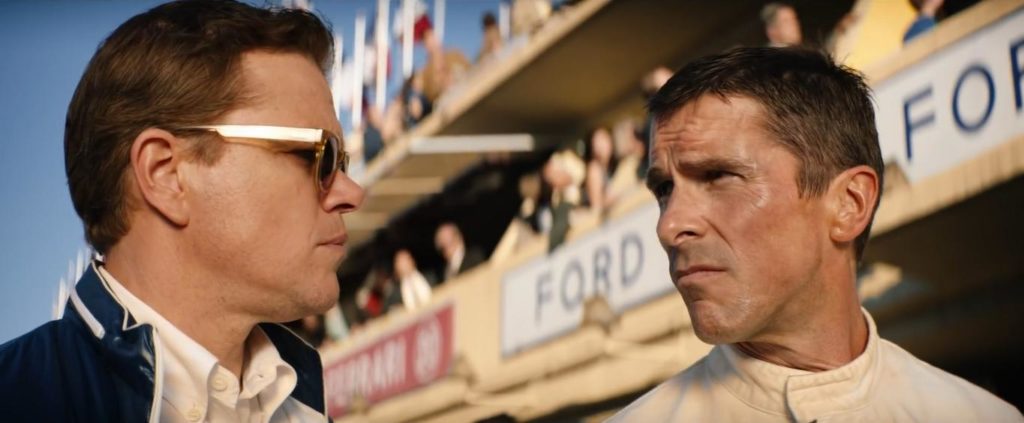 Carrol Shelby à esquerda e Ken Miles à direita se encaram num super close de seus rostos. Eles mantém uma expresão séria. Ao fundo identificamos placas com o nome "Ford" e o que parece o camarote do evento.