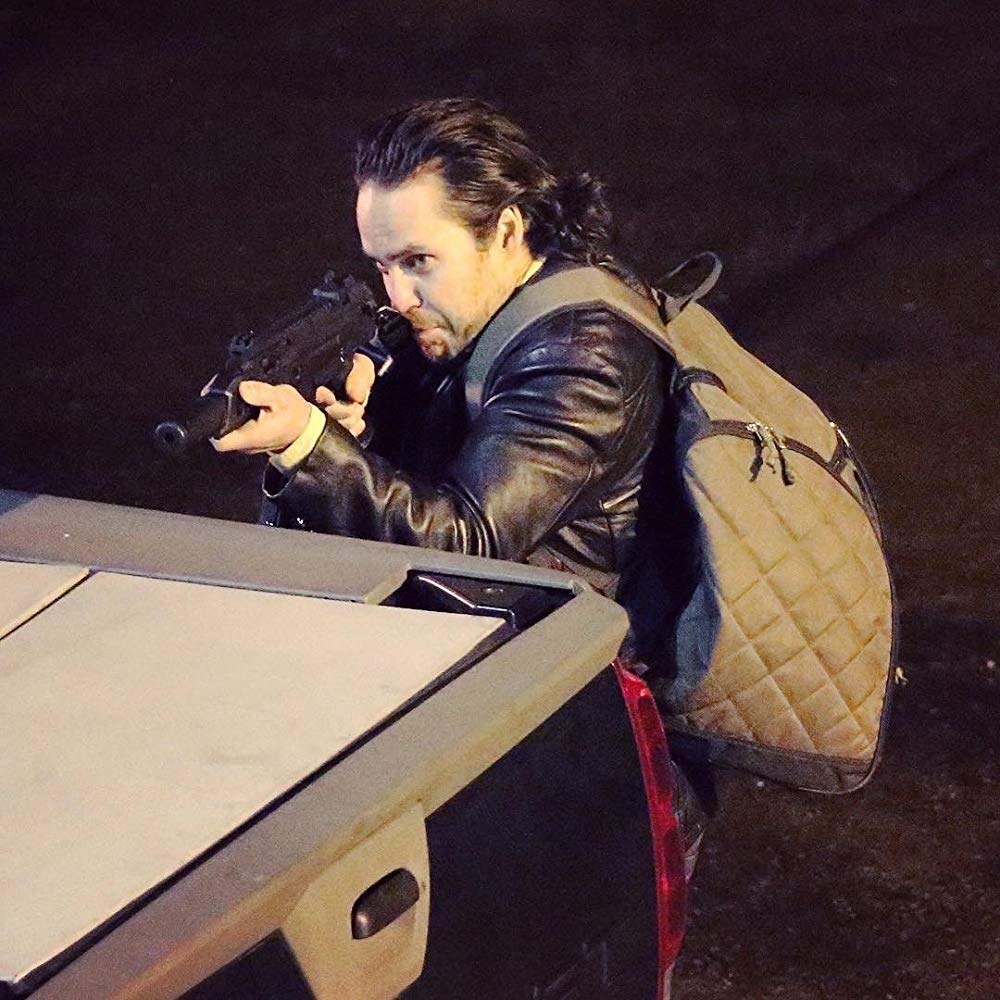 Rey, com mochila nas costas, se esconde atrás de um carro, enquanto mira com o seu rifle.
