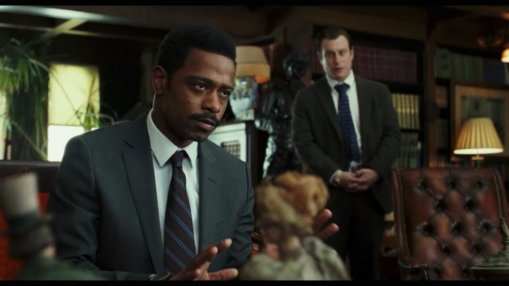 No primeiro plano, o Tenente Elliot (LaKeith Stanfield) está sentado numa poltrona, conversando com alguém (fora do quadro). Ao fundo, o policial Wagner (Noah Segan) olha o tenente
