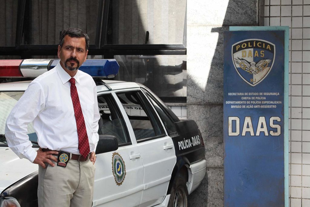 com as mãos na cintura, e distintivo no cinto, Benício (Marcos Palmeira) está próximo a uma viatura.  Na direita, está uma placa com o emblema da DAAS, dizendo "Secretaria de estado de segurança. chefa de policia. departamento de policia especializada. divisão de ação anti-seqüestro. DAAS"