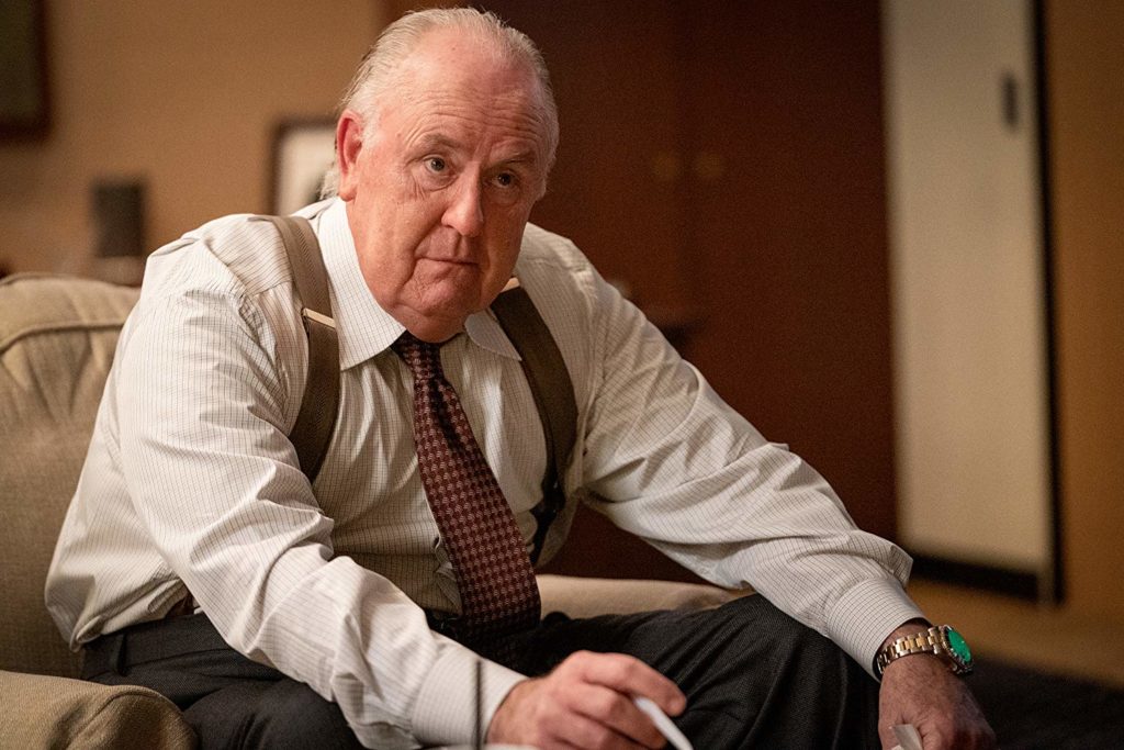 Roger (John Lithgow), sentado numa poltrona em seu escritório, segurando uma bengala