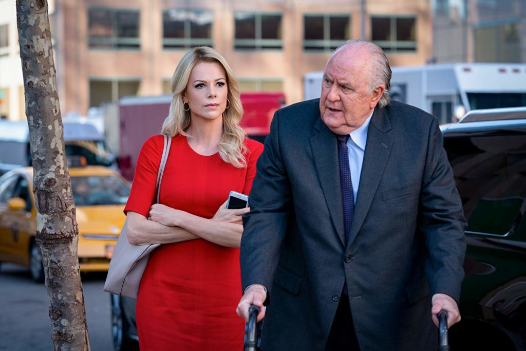 Na rua, próximo a um carro, Roger usa seu andador, e tem o olhar grave enquanto caminha. Atrás, Megyn (Charlize) está com o rosto muito grave, olhando para ele, como quem espera respostas. Ela está de braços cruzados, e segurando seu celular nas mãos.