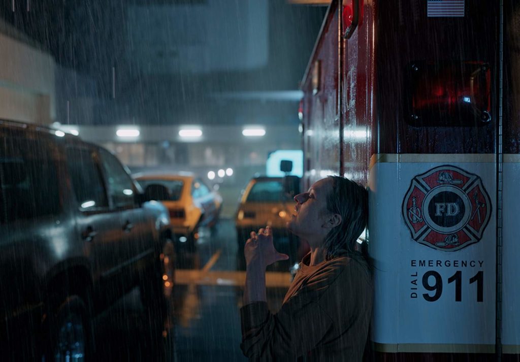 Cecilia está na chuva, apoiada numa ambulância, parada num estacionamento. Sua mão está em frente ao seu pescoço, podendo estar segurando o braço de alguém... ou não.