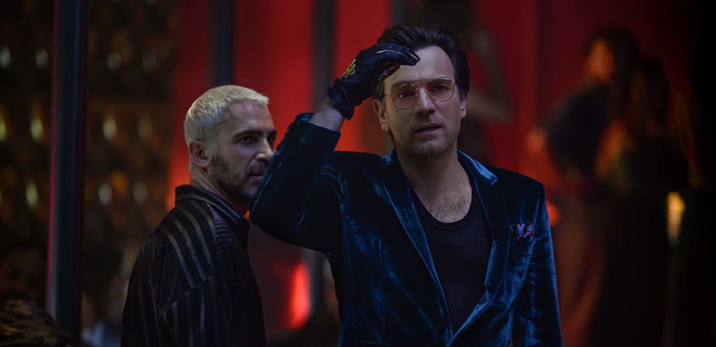 Roman sionis (Ewam McGregor) em close, usando um terno de veludo azul, luvas pretas, e óculos. Logo atrás, o Zsazs (Chris Messina)  está de camisa social listrada, de lado, olhando para Roman. 