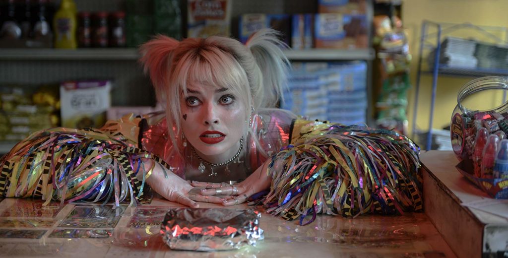 Harley (Margot Robbie) apoiando o tronco e os braços num balcão de lanchonete, com olhar choroso. à sua frente, um sanduíche embalado em papel alumínio. Ao seu lado, algumas vasilhas com doces para vender e, ao fundo, prateleiras com produtos consumíveis.