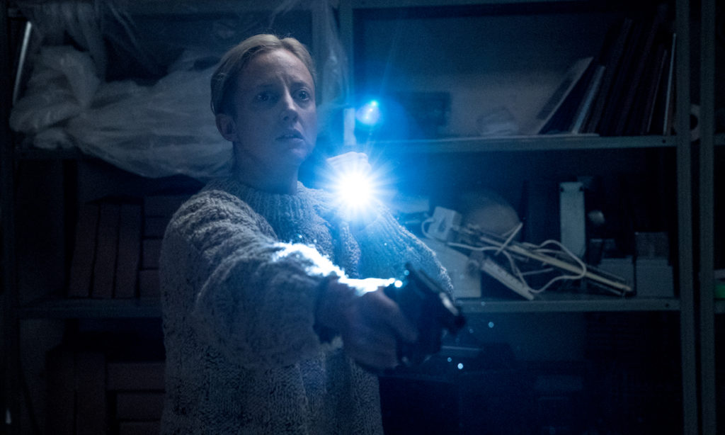 a Detetive Muldoon esta em pé, usando um pullover, com uma arma em punho na mão direita, e lanterna na mão esquerda. Ela está num almoxarifado, cuja unica fonte de luz é a própria lanterna.