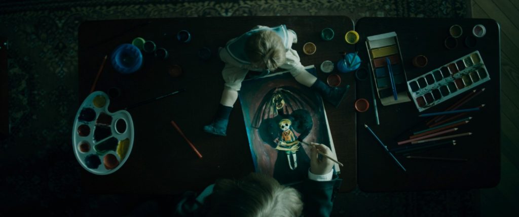 Artyom está desenhando uma figura assustadora de uma mulher abraçando uma boneca (ou uma criança). Na sua frente, um boneco de criança está olhando para a ilustração
