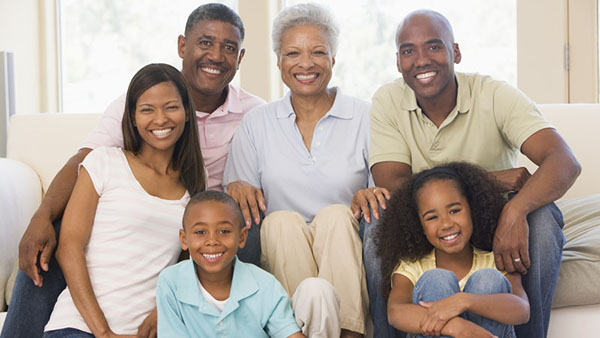 Uma família, composta por avô, avó, pai, mãe, filho e filha, sentados num sofá / no chão. Estão todos muito felizes