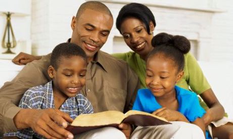 Família com pai, mãe, um menino de 4 anos e uma menina de 5, sentados juntos num sofá, lendo um livro