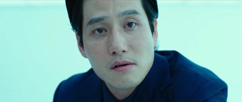 O personagem de Hae-Joon Park, sentado, olhando para a câmera. Aparenta está entediado, mas incrédulo. Ele está numa sala azul piscina