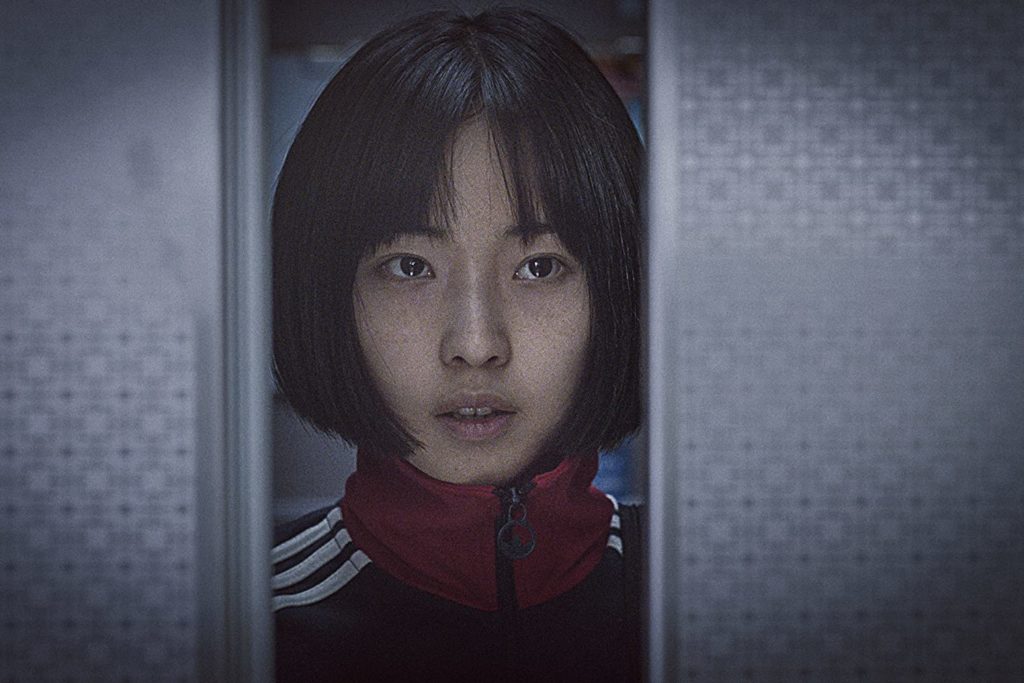 em close, Mi-na (So-nee Jeon)é vista refletida num espelho, usando sua jaqueta esportiva. A parede tem padrões repetidos, como os de um azulejo de banheiro.