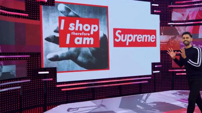 Hasan está entrando no enquadramento, mãos como se segurasse uma bola de basquete em frente ao corpo. No telão , lemos "I shop therefore I am", e a marca "Supreme"