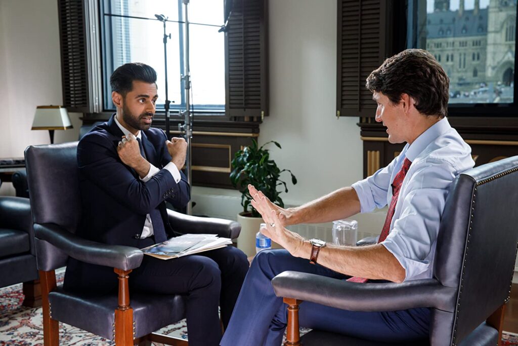 Hasan e Trudeau estão sentados num escritório com grandes janelas, com folhas de madeira abertas. Hasan está à esquerda da imagem, e podemos vê-lo de frente, em ângulo. Trudeau está mais à direita, e vemos pouco do seu rosto. Ambos estão conversando e gesticulando. Hasan faz um gesto de cruzar os braços em frente ao tronco, como quem faz o cumprimento de Pantera Negra (Wakanda Forever)