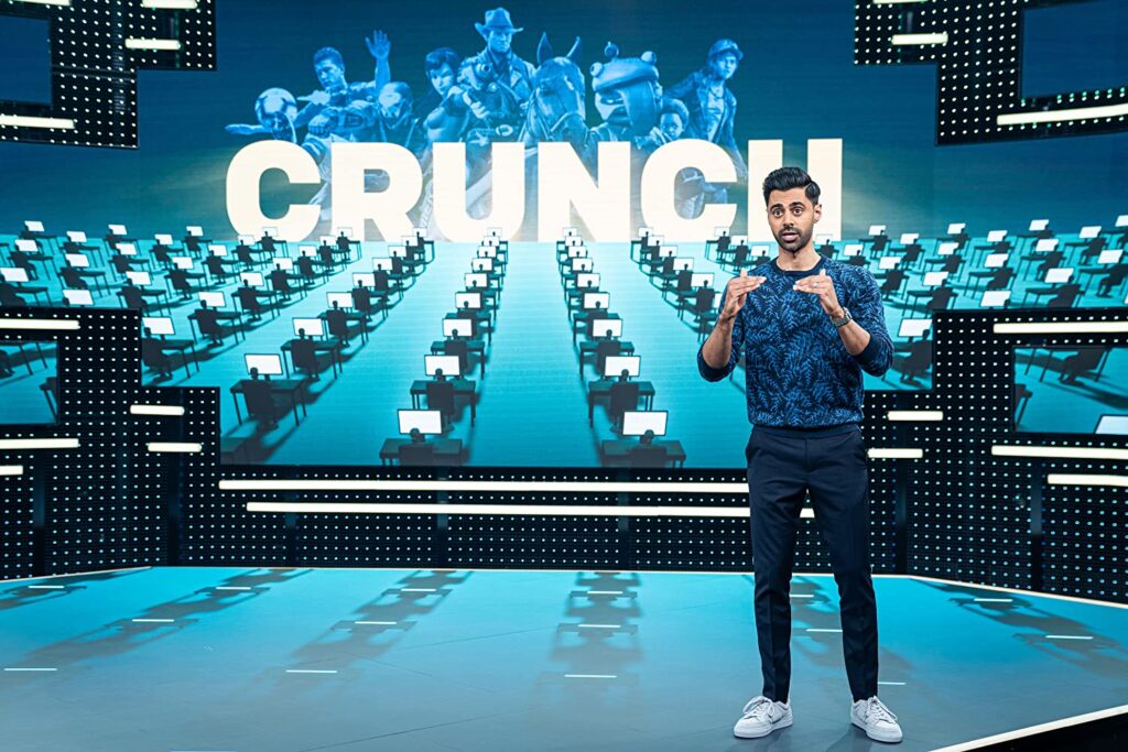 Em primeiro plano, Hasan explica um conceito. Está em pé, usando jeans e camiseta de manga comprida. Atrás, no telão, aparece em destaque a palavra "crunch". Abaixo da palavra, várias pessoas sentadas em escrivaninhas, e com telas em suas frentes. Acima da palavra, vários personagens de games famosos, como fifa, red dead redemption, overwatch e afins