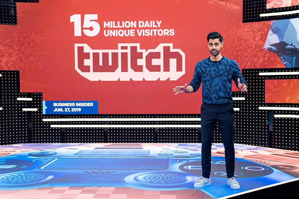 Hasan, em primeiro plano, como na foto acima, está com os braços flexionados para frente, e as mãos abertas, explicando algo. Ao fundo, lê-se "15 million daily unique visitors", e "TWITCH" em destaque. No telão, abaixo À esquerda, lemos "Business Insider, jun, 27, 2019"