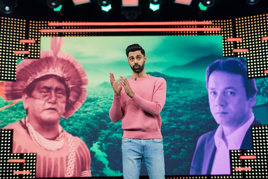 Hasan está no centro do palco. Suas mãos estão próximas, com as palmas pouco afastadas, e os dedos em riste apontando para sua latera, como se ele estivesse explicando algo. Atrás dele, no telão, vemos, na esquerda, um homem de população originária brasileiro, e do outro, um empresário.