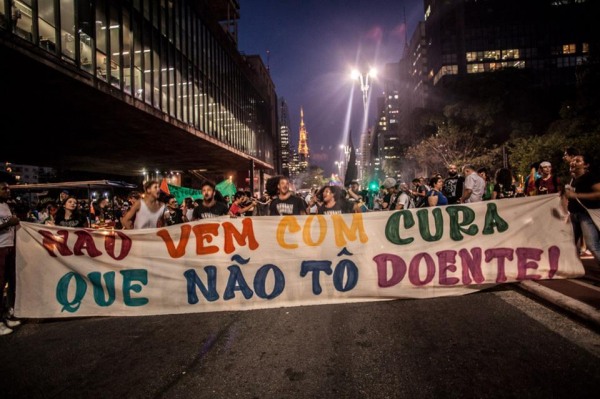 Protesto em São Paulo. À noite, diversas pessoas estão em protesto na rua. Vemos essa manifestação de frente, exibindo uma faixa onde está escrito "Não vem com cura que não tô doente!". in Posicionamento em Junho/2020