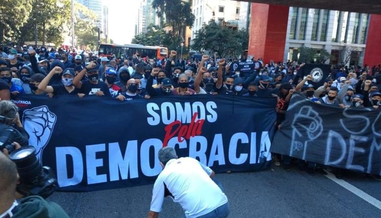 Movimento pela democracia, em São Paulo. Uma multidão de pessoas, majoritariamente usando preto e máscaras faciais, protestando e com braços erguidos. Na frente delas, vemos uma faixa escrita "Somos pela Democracia". Existe mais uma faixa à direita da foto, mostrando um punho desenhado em linhas brancas, e há algo escrito ao seu lado, mas está ilegível. As pessoas estão sob o vão do MASP. in Posicionamento em Junho/2020