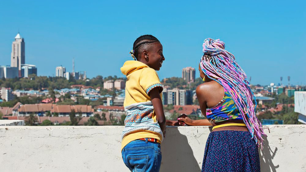 Kena e Ziki conversam no muro do prédio, olhando a vista da cidade de Nairobi.
