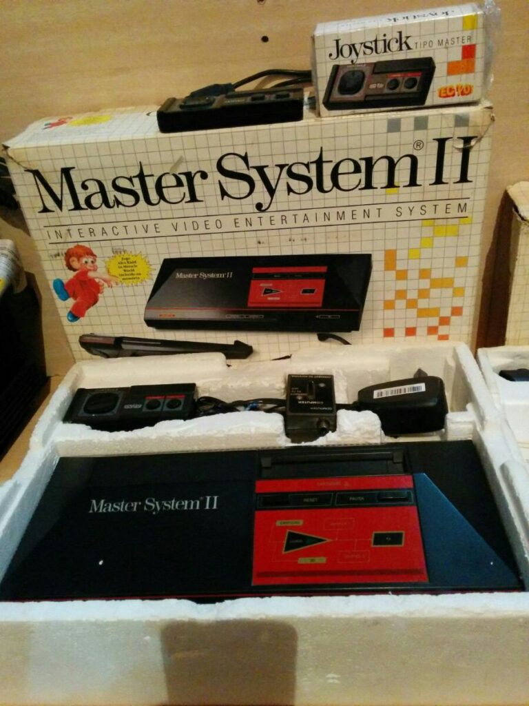 Imagem de um Master System 2 com sua caixa ao fundo, e armazenado em seu container de isopor