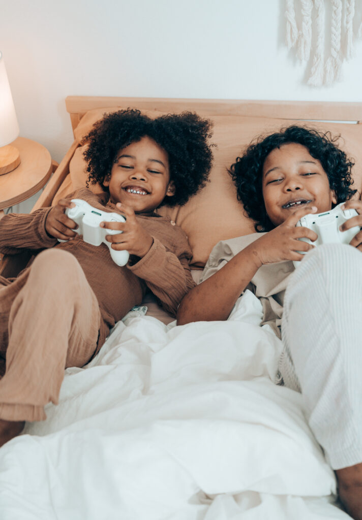 Duas crianças na cama deitadas com controles de videogame, sorrindo