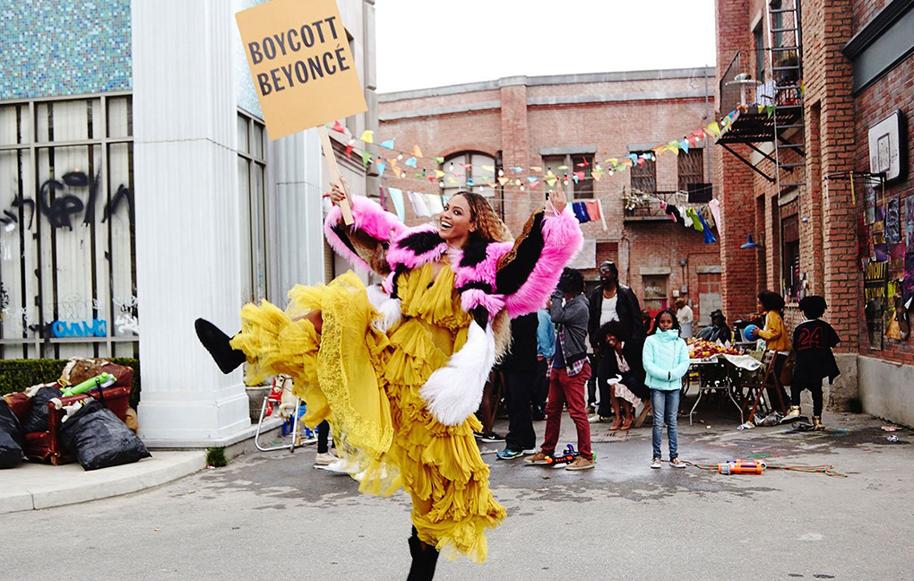 Beyoncé segura uma placa escrita "Boycott Beyoncé" (Boicote Beyoncé) enquanto dança e ri em um vestido amarelo.