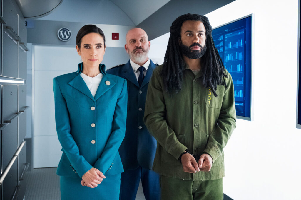 Na sala das Gavetas, Melanie (em seu uniforme de secretária) e Andre (numa roupa que lembra de um presidiário). Estão ambos com as mãos próximas, à frente do corpo (André está algemado). Atrás deles, Roche (Mike O'Malley) está esperando por algo.