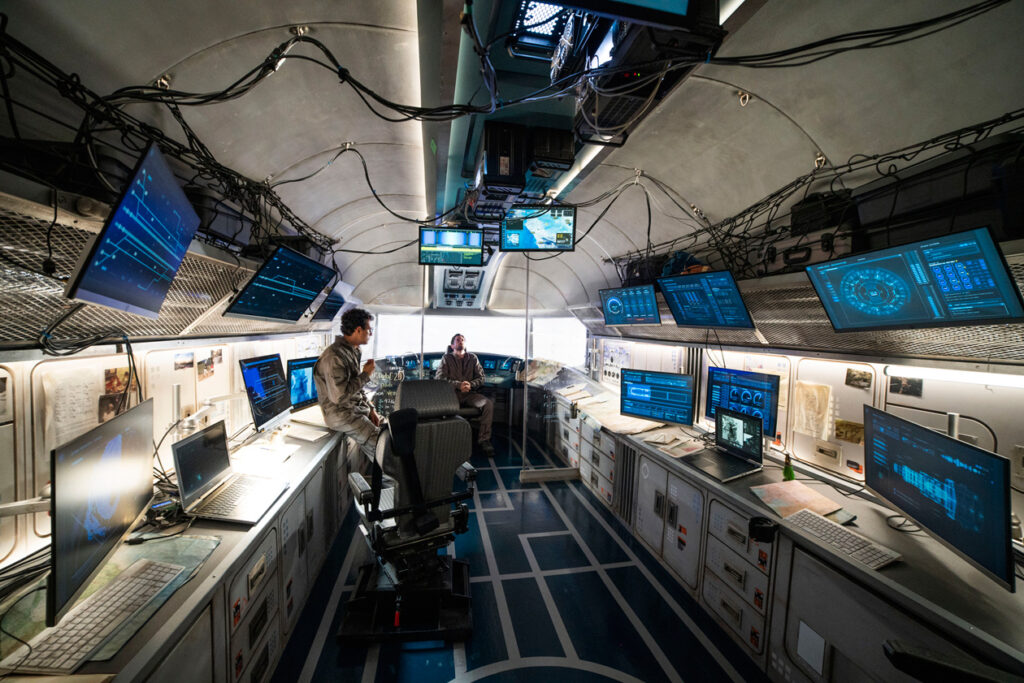 Vista da sala central de operações da Snowpiercer. A bancada está disposta em U por toda a parede, e existem vários monitores por toda a sala. É possível ver Javier e Bennet