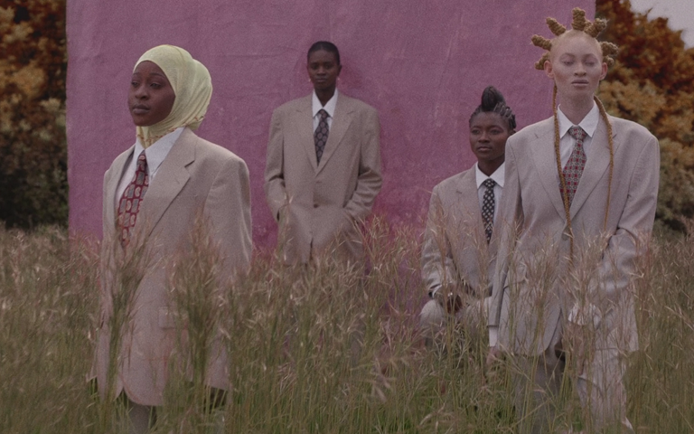 Quatro mulheres negras de terno bege num campo verde. À esquerda, uma mulher negra com os cabelos cobertos por um tecido claro, mais ao fundo uma mulher encostada em uma espécie de parede roxa, outra mulher sentada com um penteado típico africano e à extrema direita uma mulher albina.