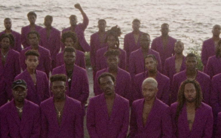Vários homens negros alinhados em 4 fileiras usando ternos roxos, ao fundo um dos homens ergue o punho em orgulho.
