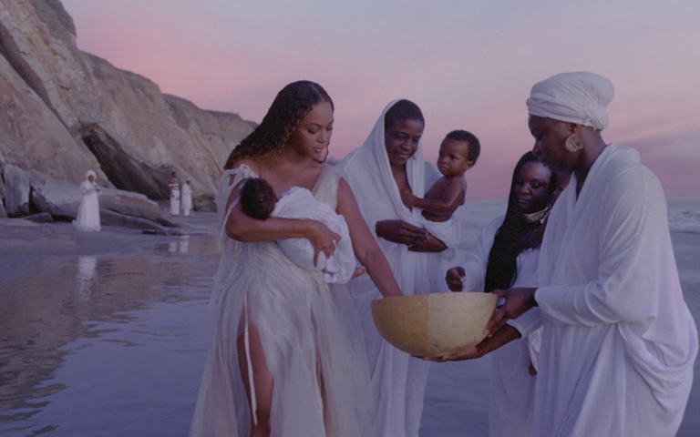 Numa praia, no fim da tarde Beyoncé e mais duas mulheres negras performa um ritual de nascimento, molhando um bebês em seus colos. Uma outra mulher negra segura uma espécie de cuia. Mais ao fundo outras pessoas negras. Todos de branco.