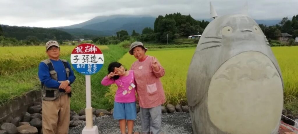 à direita, a estátua do Totoro em tamanho "real". À esquerda, o escultor (de braços cruzados), a placa de ônibus no ponto, e uma senhora idosa e uma criança (fazendo V com os dedos das mãos) posando para as fotos.
como visto em Notícias de Sofá 006 - Carrefour, estátua de Totoro, e ladrão vegetal