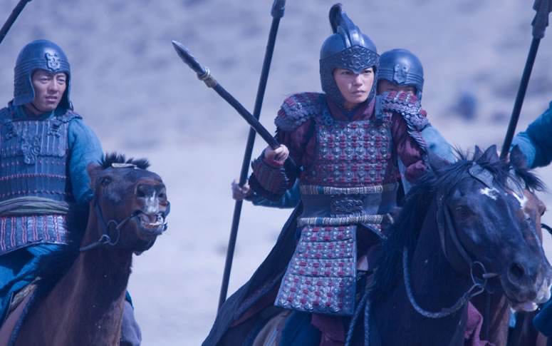 Mulan, com armadura completa, portando lança no braço direito, seguida por mais 3 cavaleiros (só vemos 2 deles), está montada em seu cavalo.