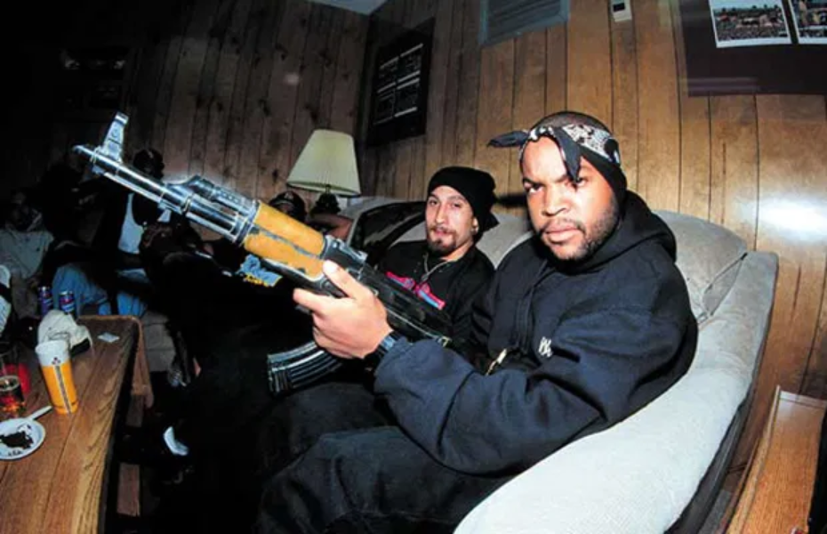 A imagem mostra Ice Cube segurando um fuzil Ak-47, com o rapper B-Real no fundo