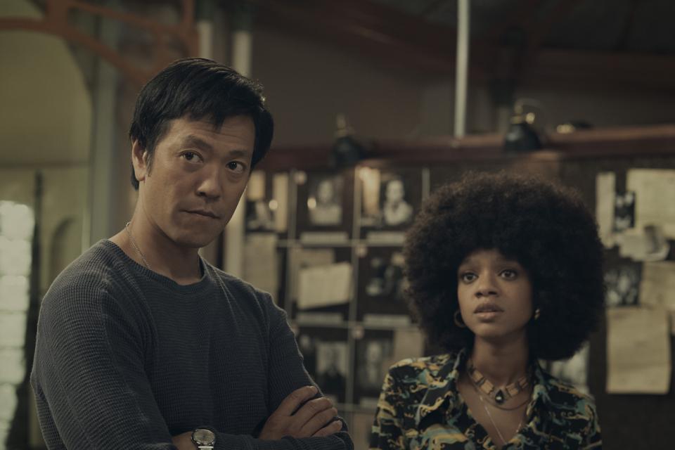 Louis Ozawa (Joe Mizushima) está com os braços cruzados e uma expressão inquisitiva ao lado de Tyffany Boone (Roxy Jones), que parece surpresa.