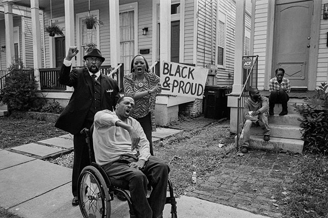 Três pessoas (um homem em pé, uma mulher e um cadeirante) protestam. A mulher segura a placa "Black and Proud".