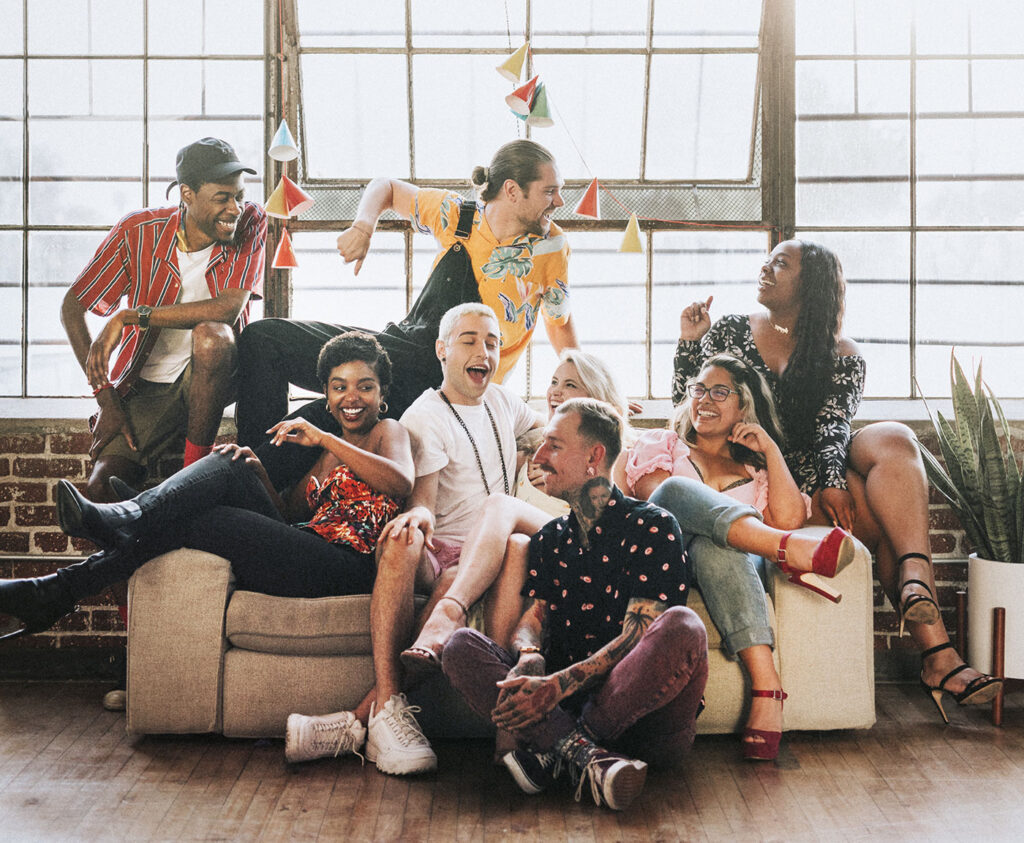 Grupo de jovens adultos, 4 homens e 4 mulheres, 3 negros, 2 brancos, 2 mestiços, com visual alternativo, sentados no sofá ou no chão, rindo e se divertindo