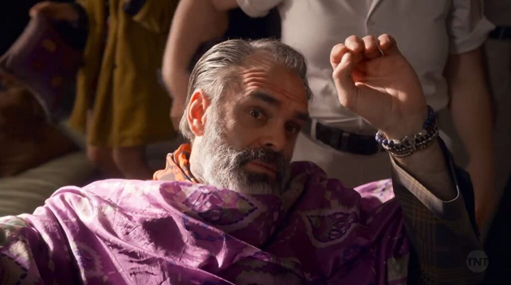 Pike usando camisa social e relógios, além do cabelo com gel. Está observando alguém com atenção despretensiosa