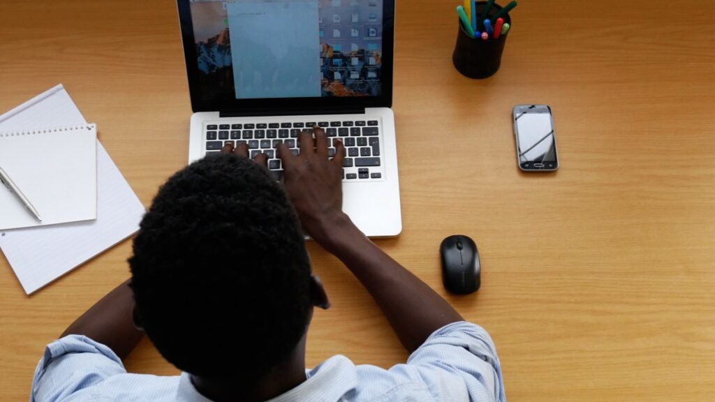 Fotografia de cima de um homem negro utilizando um laptop (um Mac), com mouse sem fio, celular, copo com lápis e blocos de papel próximos. A mesa é de madeira clara, e bem limpa (tudo parece caro).
