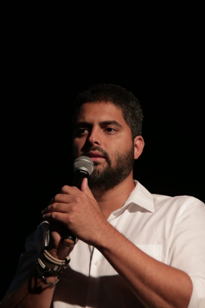 Hugo Canuto, segurando microfone em mãos, num fundo preto (provavelmente dando palestra num teatro)