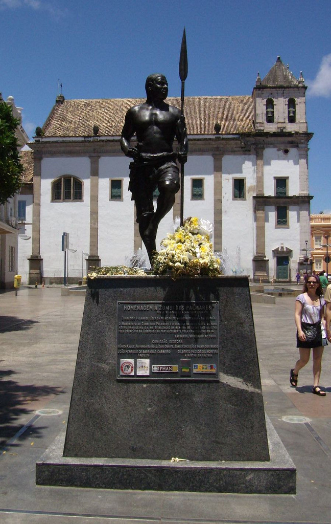 Estatua de Zumbi de Palmares, de material preto, ao fundo vemos a catedral de salvador.